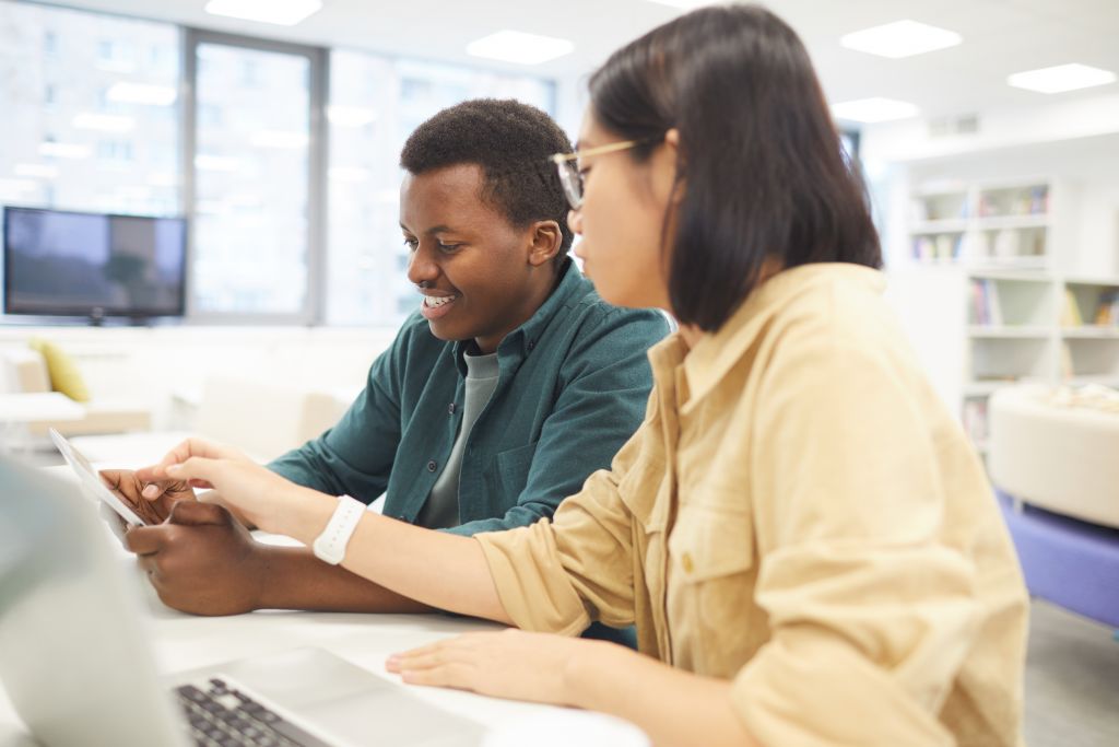 Portrait of two students studying in college library, focus on Asian young woman using laptop in foreground.