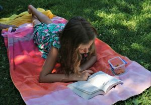 Girl laying on a blanket, outside, and reading a book.