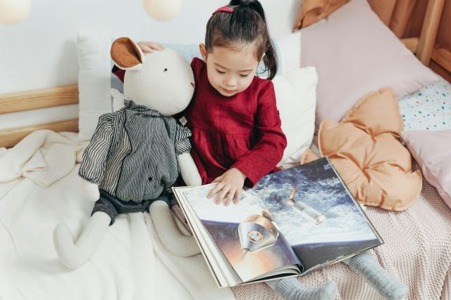 Little girl sitting on her bed with stuffed animals and reading a book.