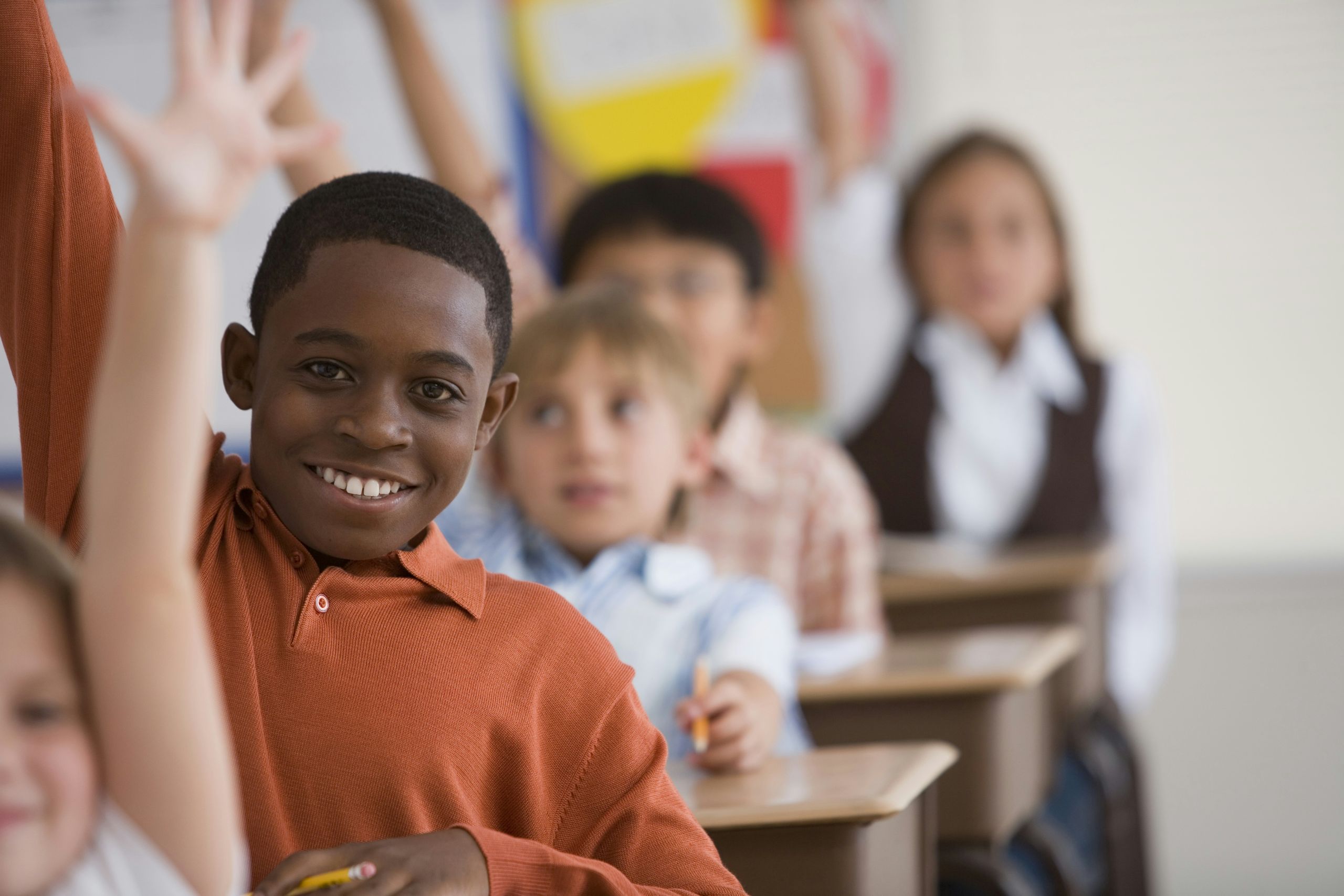 Young students holding up their hand, smiling, in class.