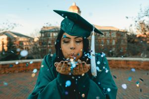 A person in their graduation cap and gown, blowing confetti.