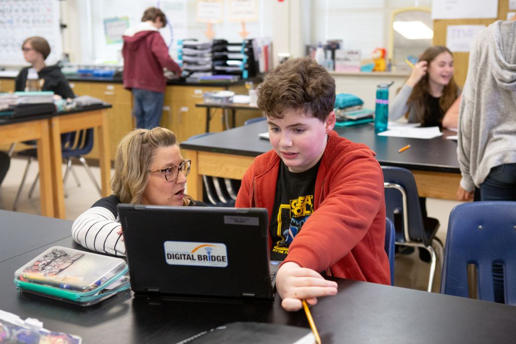 A teacher kneeling near a student's desk to talk to the student