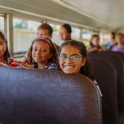 Three elementary girls smiling as they peek over school bus seat