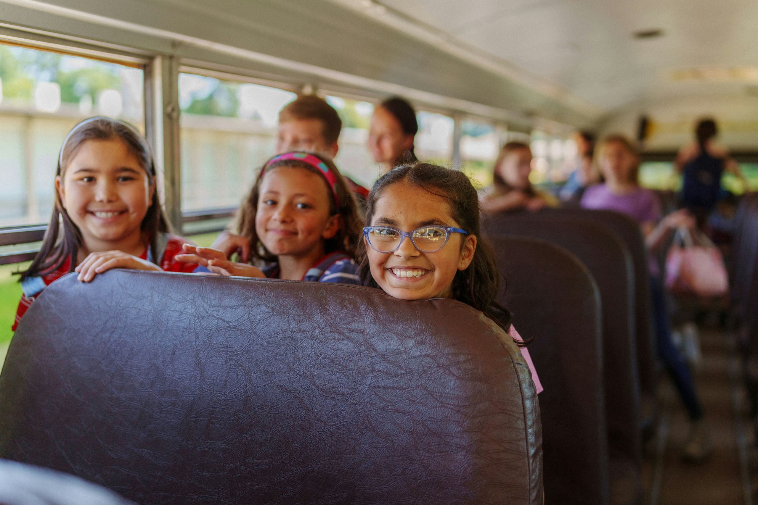 Three elementary girls smiling as they peek over school bus seat