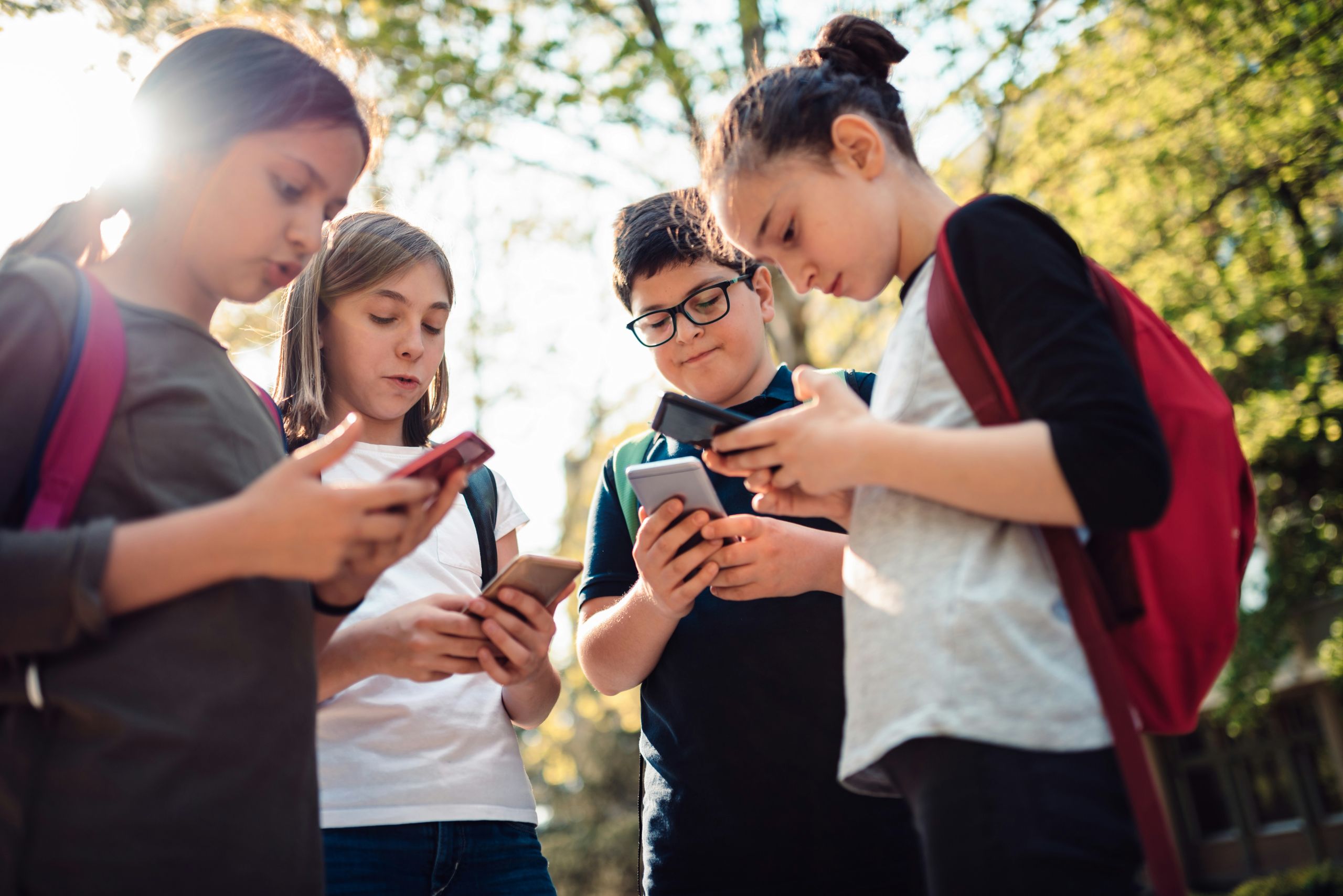 getty-images-xQHXLEfMJg0-unsplash A group of middle school students each with a phone in their hands and eyes towards screens. They are outside with trees around them.