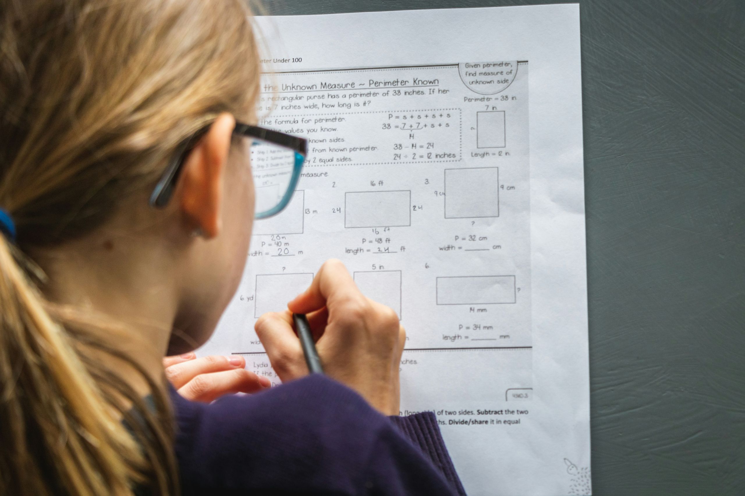 greg-rosenke-DJWvzY4pkGU-unsplash A girl with glasses & blonde hair working on a worksheet taped to a chalkboard
